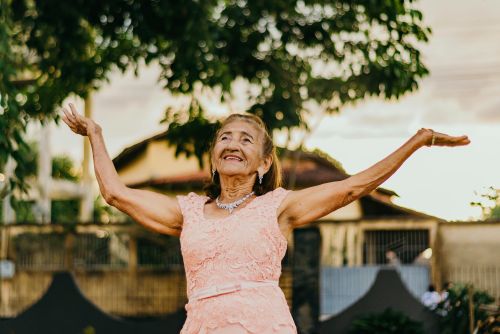 Elderly woman in a pink lace dress smiling and posing outdoors.